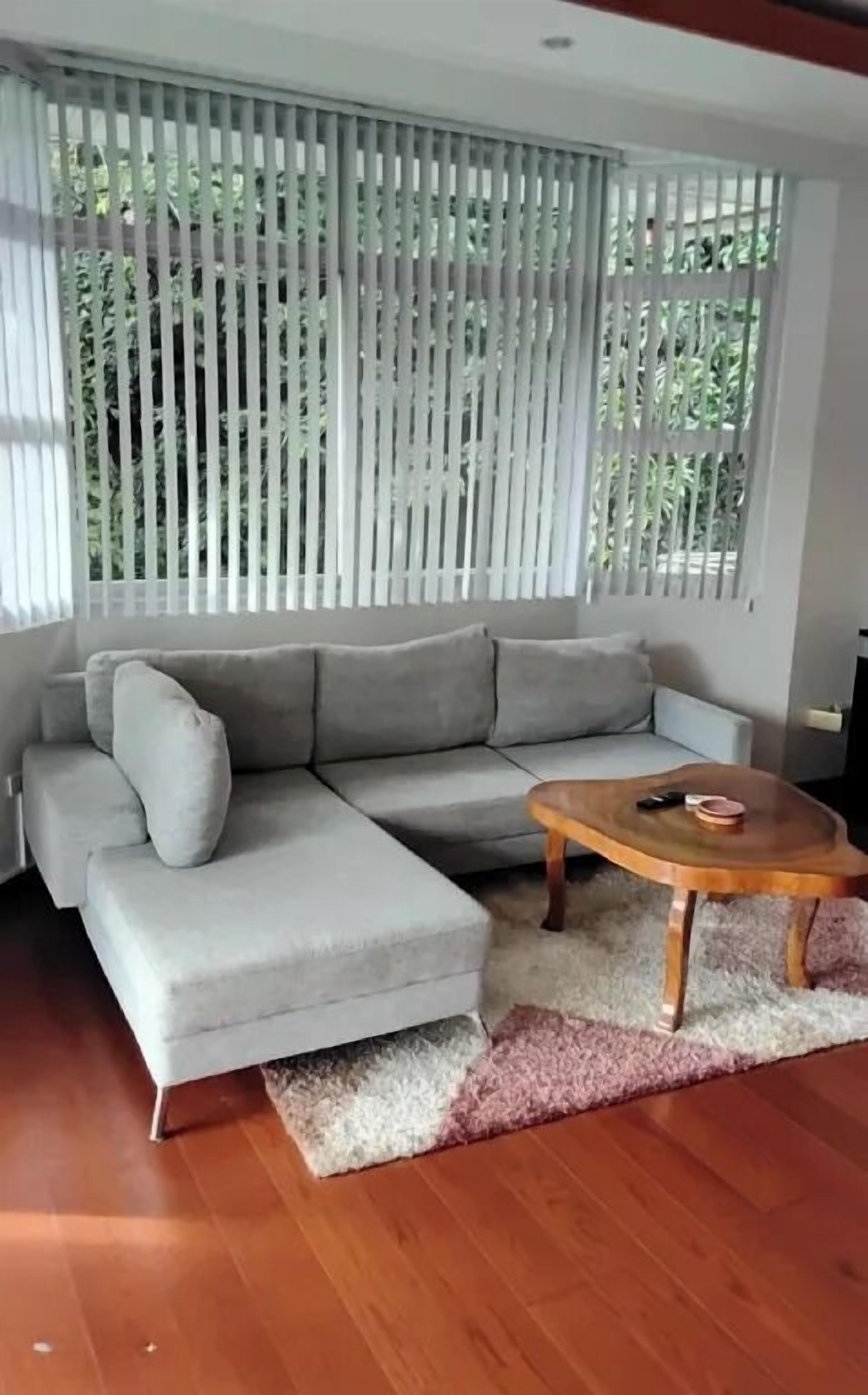 Een moderne woonkamer met een grijze modulaire bank, een houten salontafel en een groot raam met verticale blinds.
