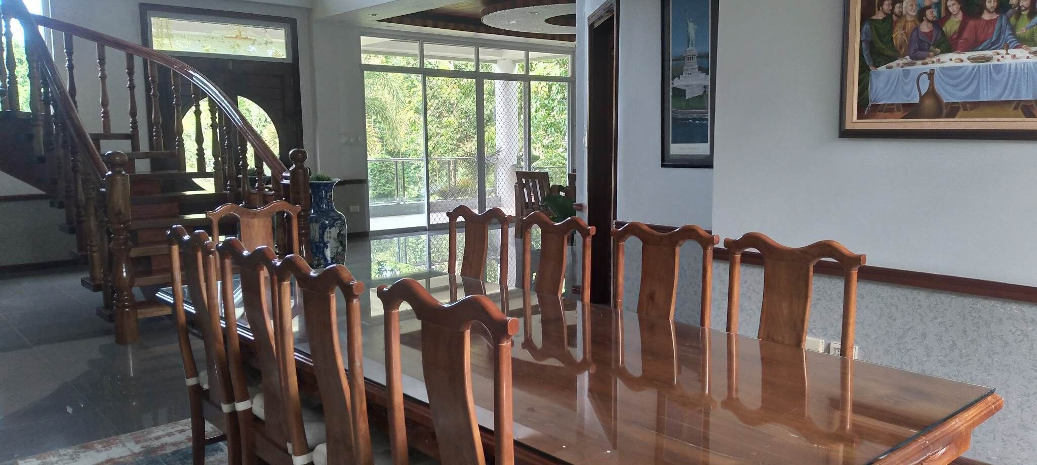 A dining area with a long wooden table and chairs, a staircase, and a large window with a view of greenery.