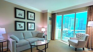 Habitación de hotel con sofá, sillón y una mesa de centro de vidrio. Se observan cuadros en la pared y una vista de la ciudad a través de amplios ventanales.