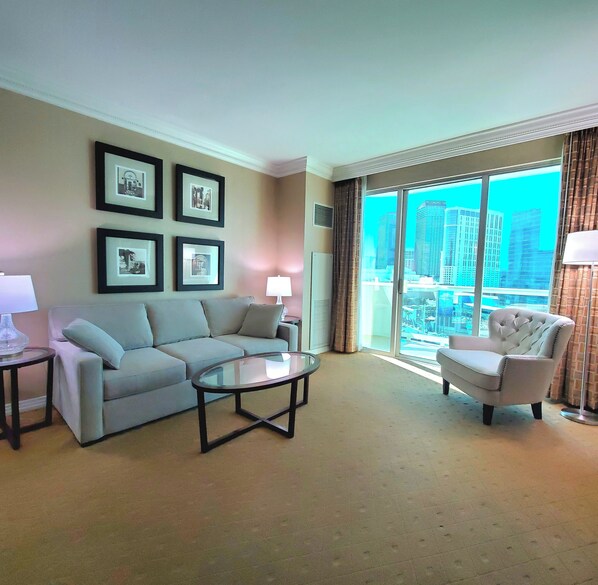 Habitación de hotel con sofá, sillón y una mesa de centro de vidrio. Se observan cuadros en la pared y una vista de la ciudad a través de amplios ventanales.