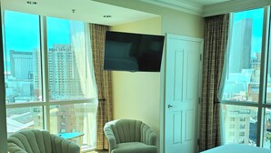 Habitación de hotel con una cama grande, dos sillones, televisor en la pared y un balcón con vista a la ciudad.
