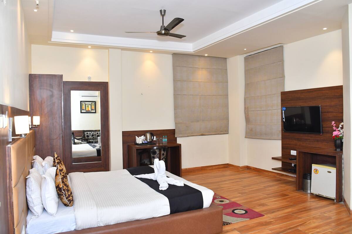 A hotel room with a bed, a TV, a ceiling fan, a wooden headboard, a mirror, a small table, and a window with blinds.