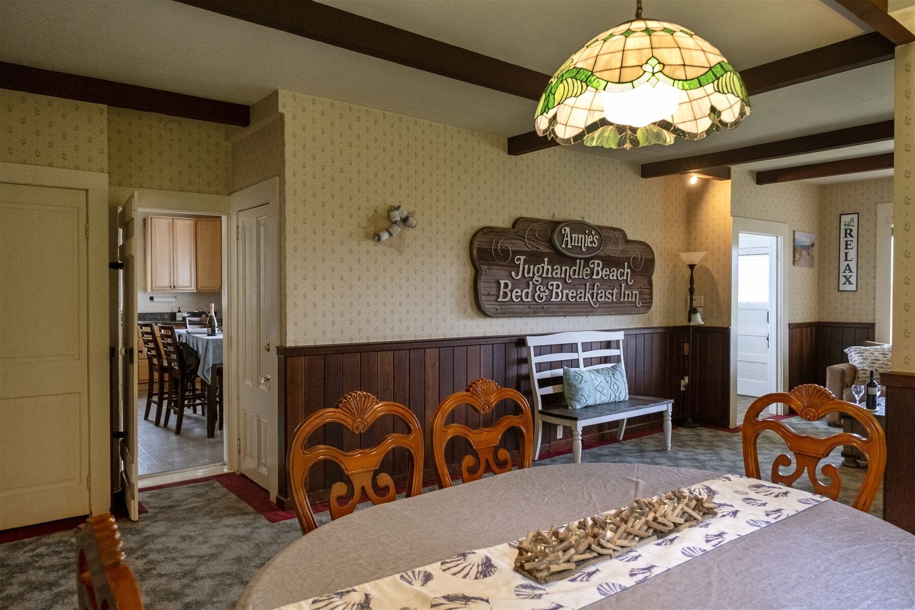 A cozy room with a bed, a bench, and a table with a decorative centerpiece. A sign on the wall reads "Annie's Tughandle Bench Bed & Breakfast Inn."