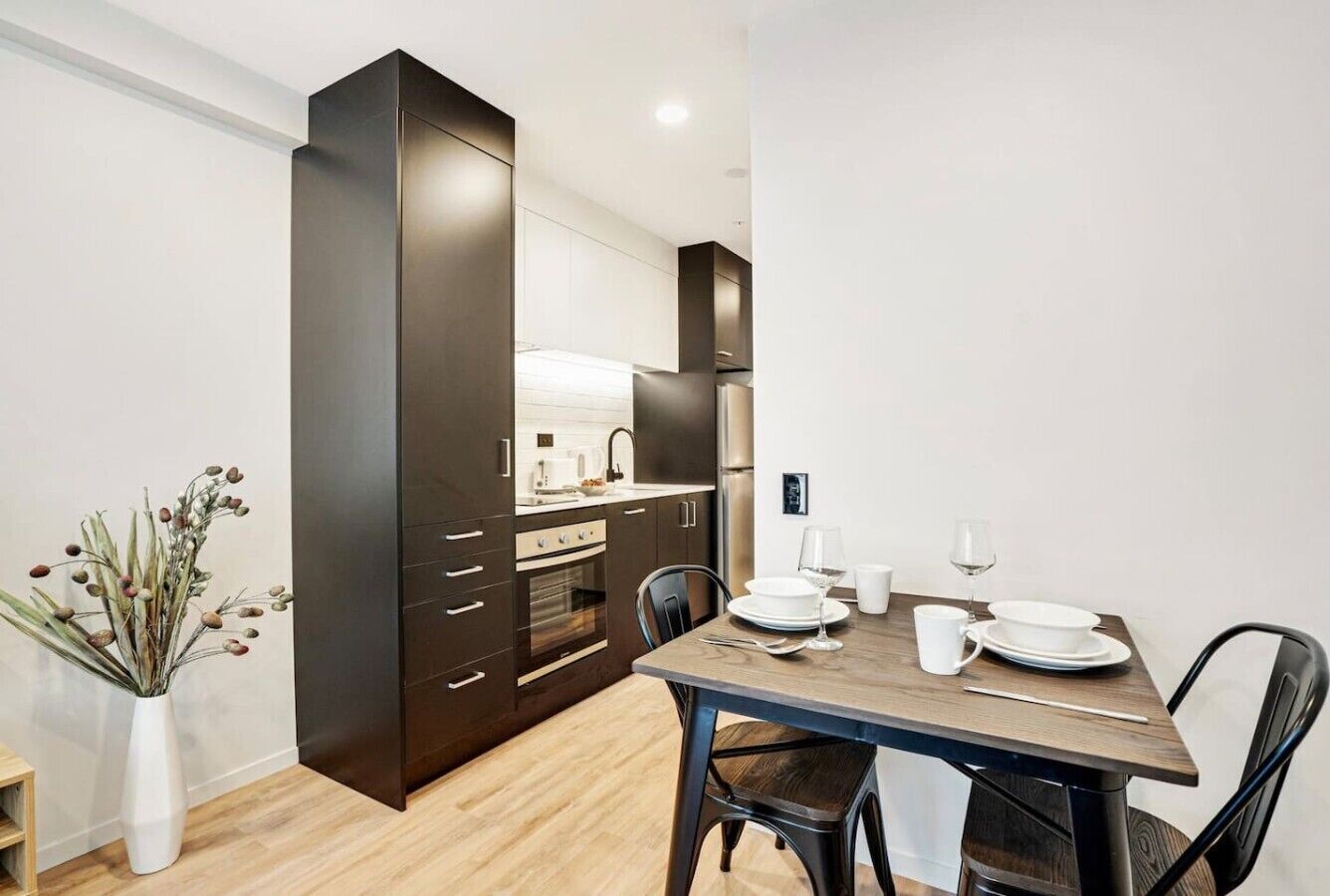 A modern kitchen with dark cabinetry, a dining table with chairs, and a vase with dried flowers.