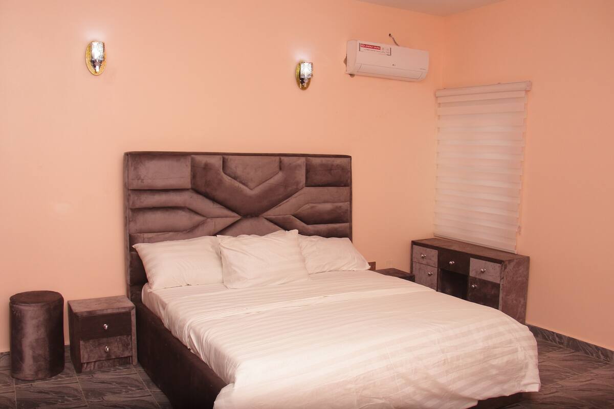 A bedroom with a large bed, two wall-mounted lights, an air conditioner, a bedside table, and a window with blinds.