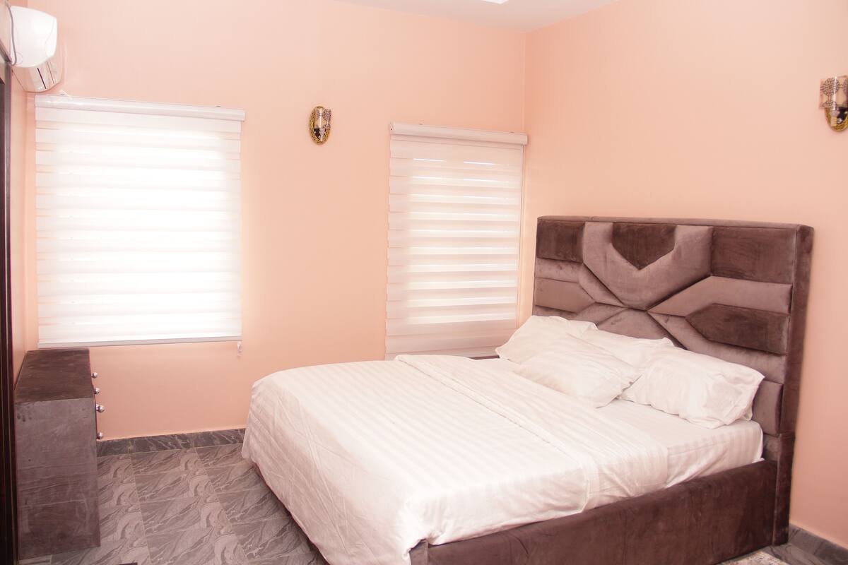 A bedroom with a bed, two windows with blinds, a wall-mounted light fixture, and a dark-colored dresser.
