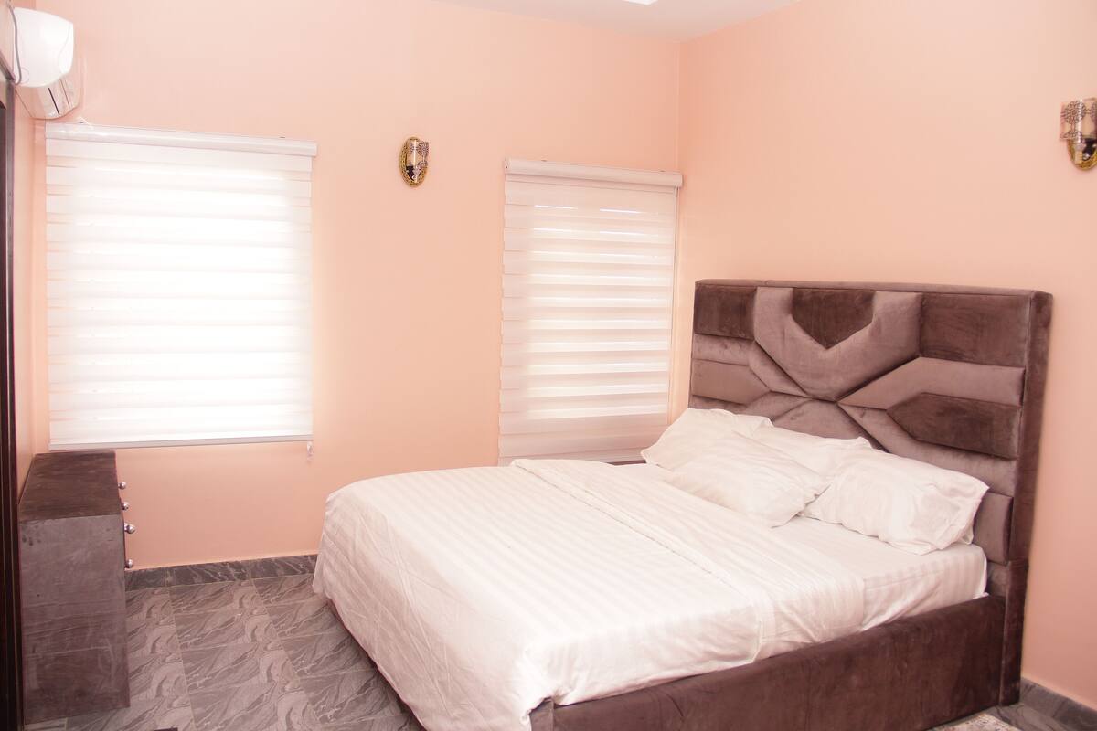 A bedroom with a bed, two windows with blinds, a wall-mounted light fixture, and a dark-colored dresser.