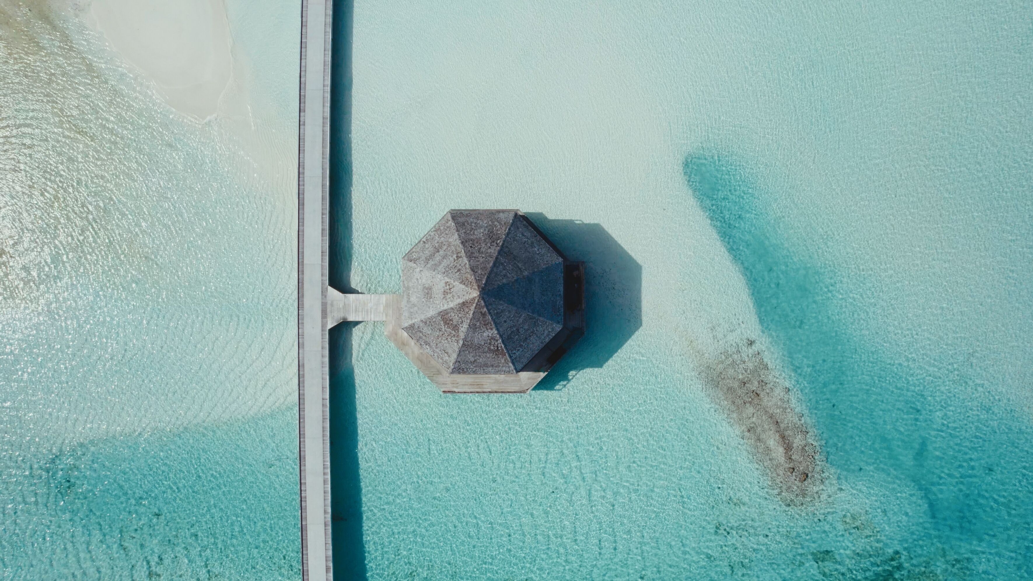An aerial view of a beach with a hexagonal structure, possibly a gazebo or shelter, near the water's edge.