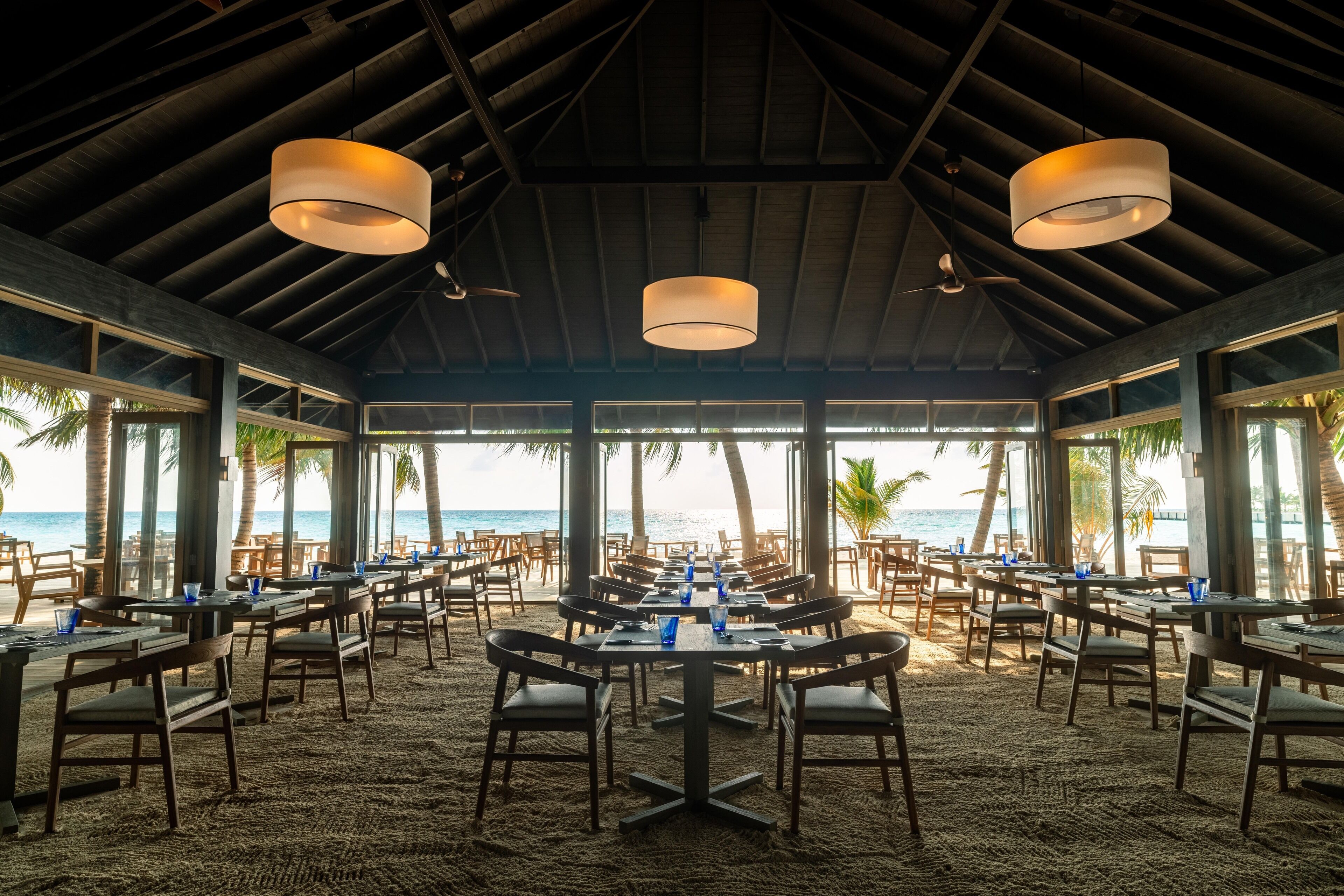 Un restaurante frente a la playa con mesas y sillas dispuestas sobre arena, amplios ventanales con vistas al océano y un techo alto con vigas a la vista.