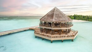 Un resort sobre el agua con varios niveles, techo de paja, múltiples zonas de comedor y una terraza de madera.