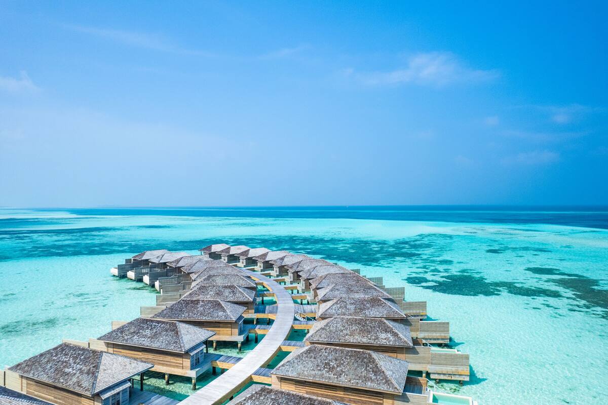 Ett flygbildsfoto av bungalows som är byggda över vattnet, med en tydlig gångväg som leder till dem, omgivna av turkosblått vatten och en klar himmel.