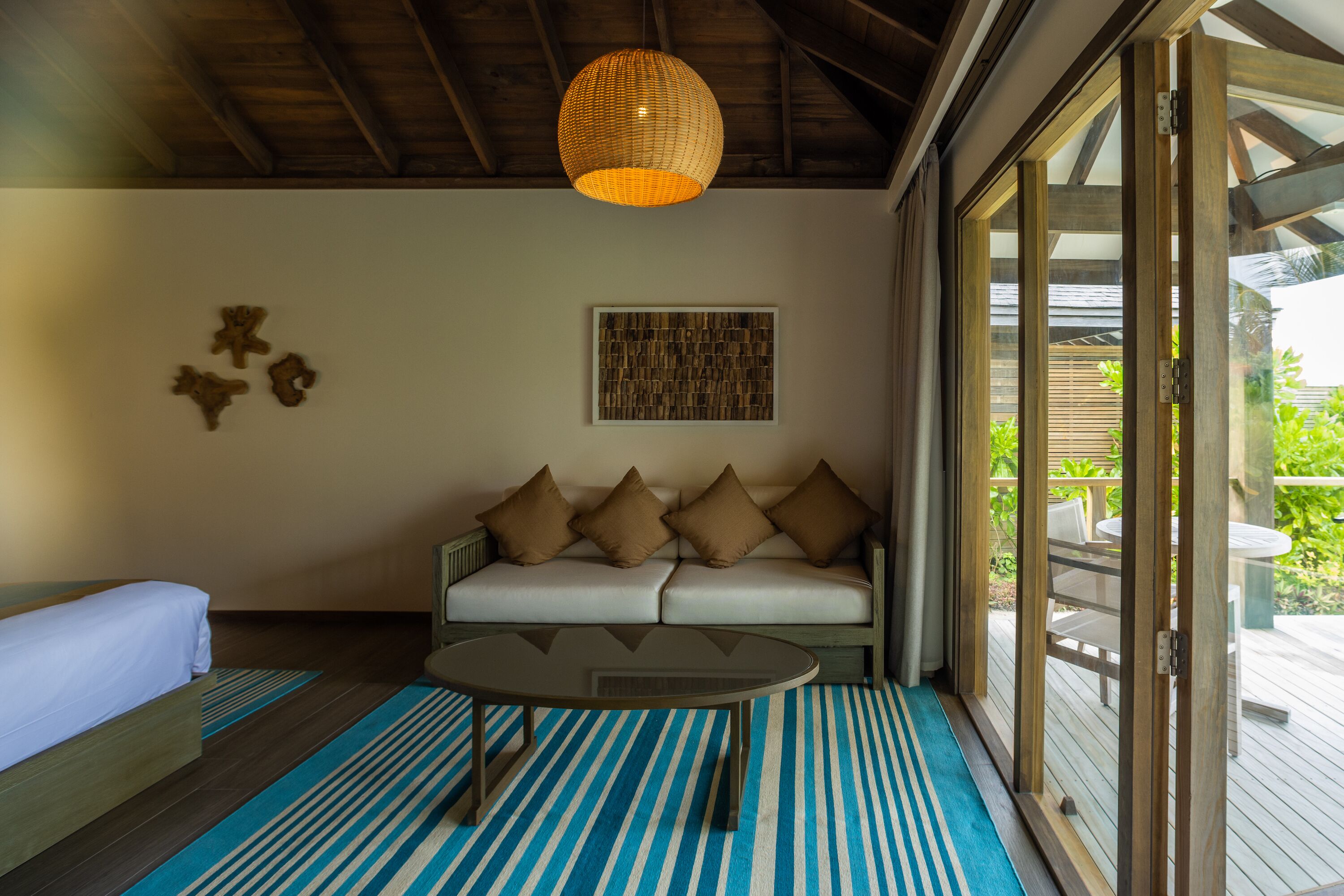 A room with a bed, a sofa with cushions, a coffee table, a hanging light fixture, and a large window with a view of greenery.