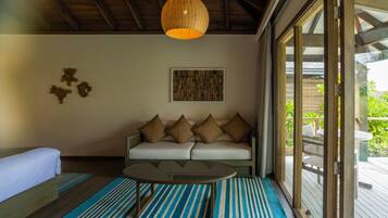 Una habitación con cama, sofá con cojines, mesa de centro, lámpara colgante y un amplio ventanal con vista a zonas verdes.