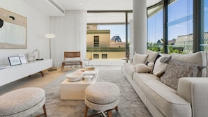 Un salon moderne comprenant un canapé, un pouf, une table basse, une table d’appoint et un fauteuil. Une grande fenêtre offre une vue sur les bâtiments environnants.