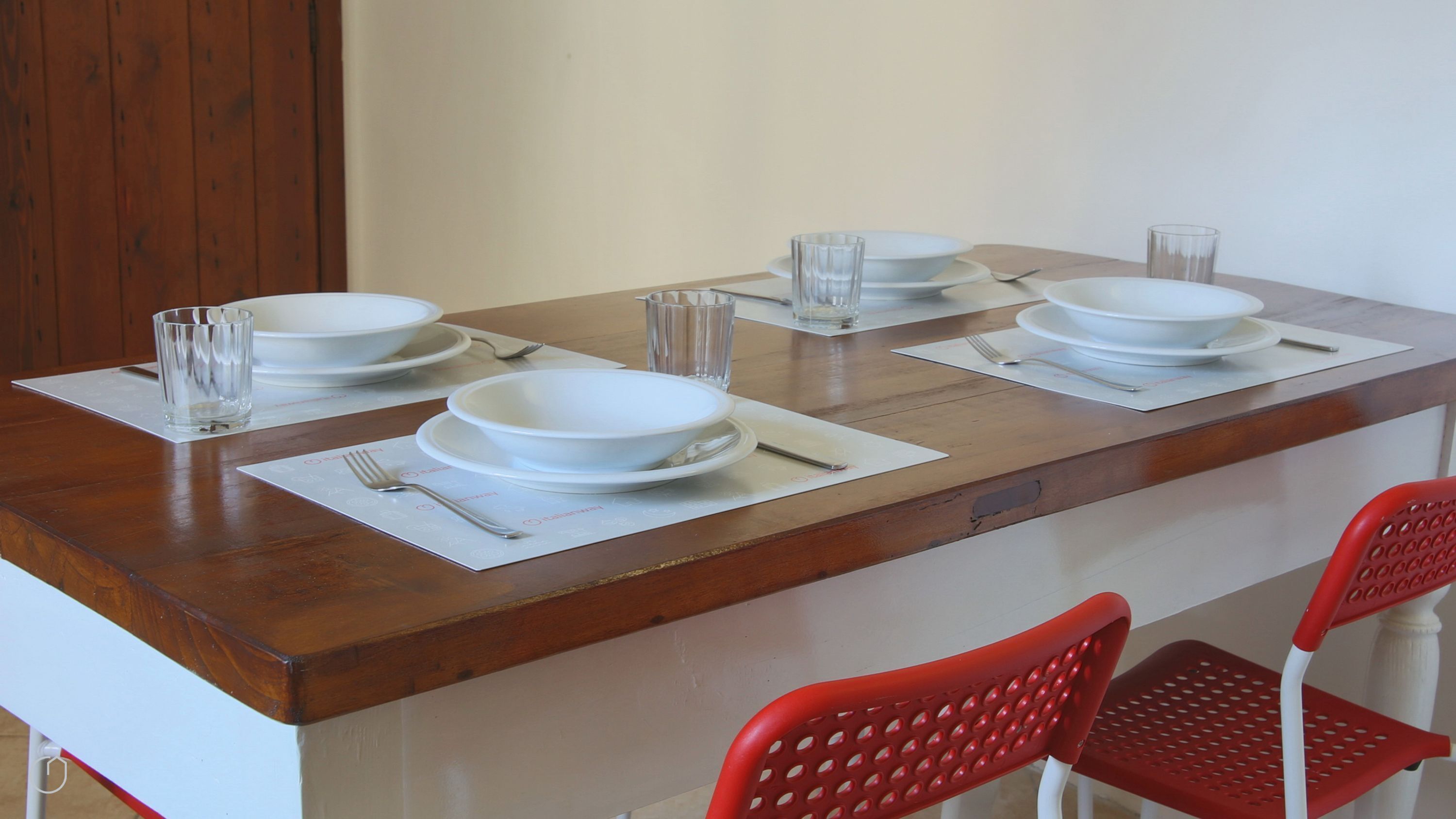 Una mesa de comedor dispuesta con platos blancos, vasos y cubiertos, acompañada de sillas rojas.