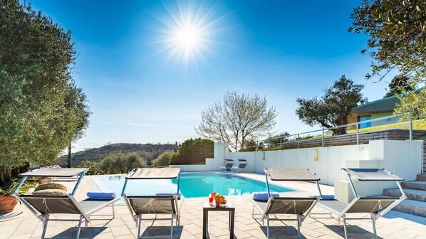 A poolside area with sun loungers, a table with a drink, and a view of the pool and hills.