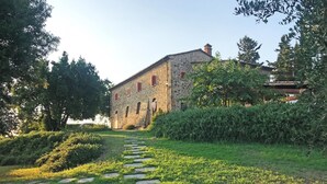 A stone building with arched windows and a red door, surrounded by lush greenery and a stone pathway. - Cosciano 12 Emma Villas (Pomarance)