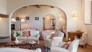 A cozy living room with a sofa, armchairs, a coffee table, and a desk with a chair. There is a bookshelf and a lamp. - Cosciano 12 Emma Villas (Pomarance)