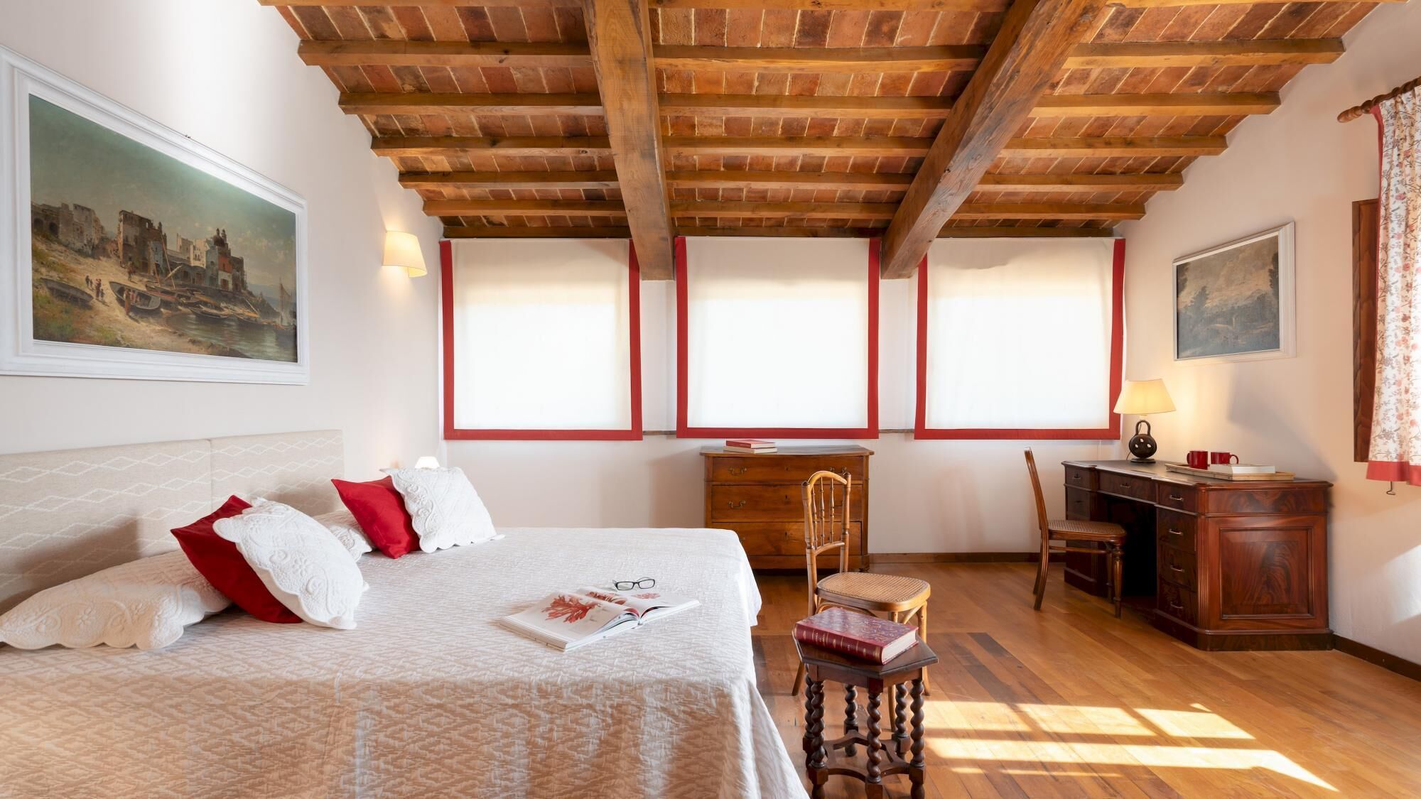 A bedroom with a bed, a desk, and a chair. There are windows with blinds, a painting on the wall, and a lamp on the desk.