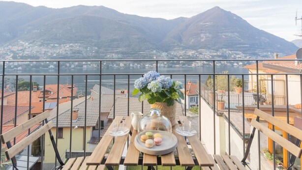 A balcony with a table set for tea, a vase with flowers, and a view of a lake and mountains. - Pozzo Vertical Apartment With Garden and Parking (Torno)