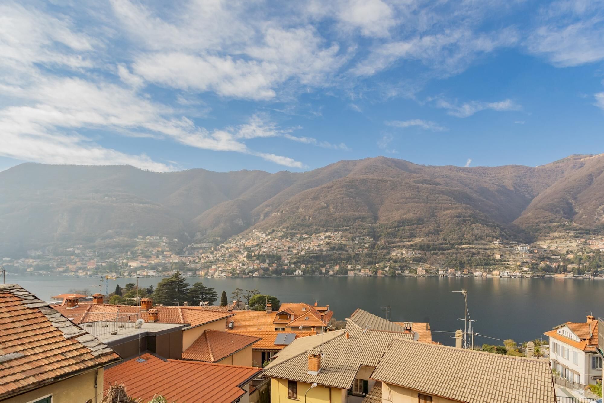 A town with terracotta-roofed buildings, a lake, and mountains in the background.