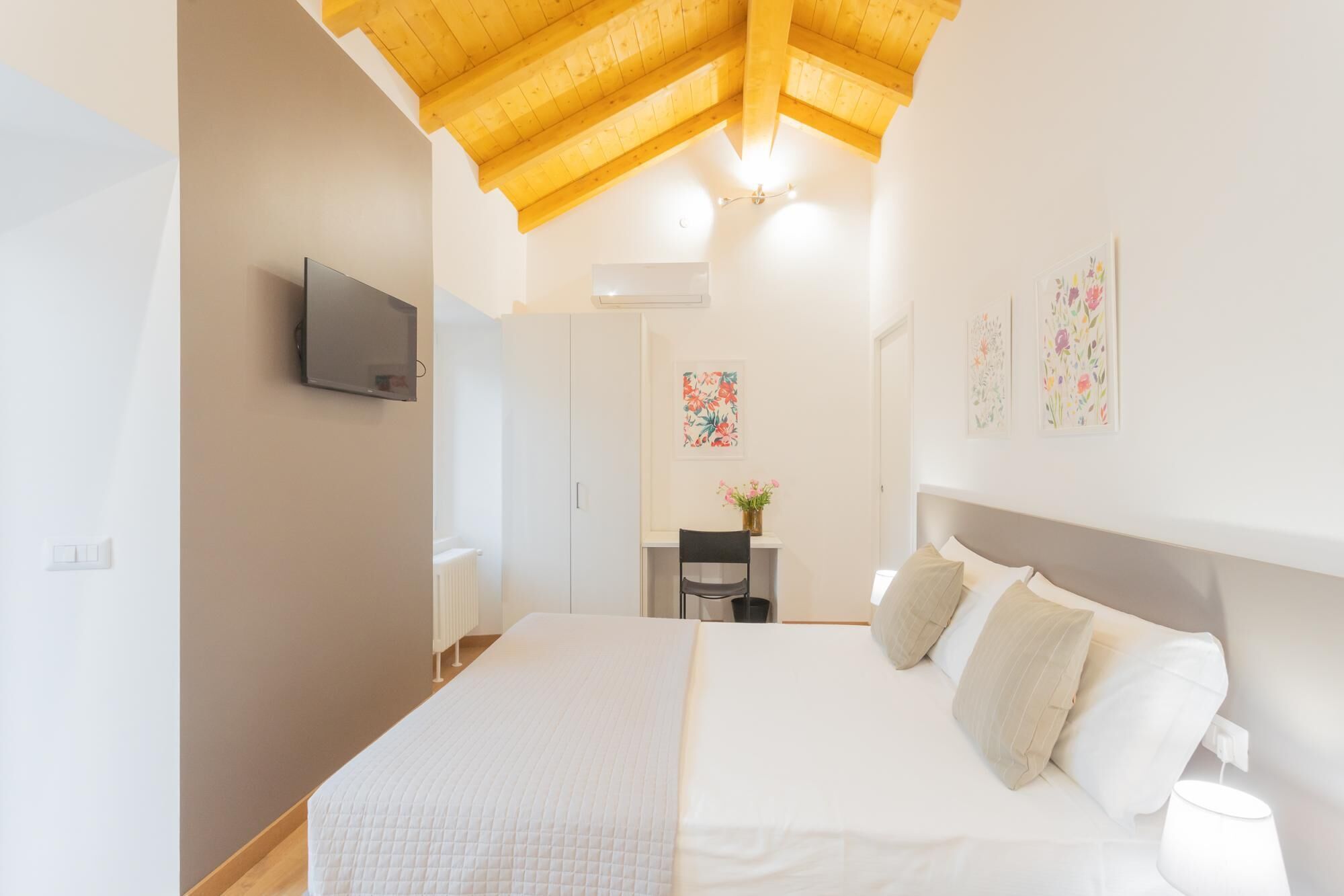A bedroom with a large bed, a TV mounted on the wall, and a ceiling with wooden beams.