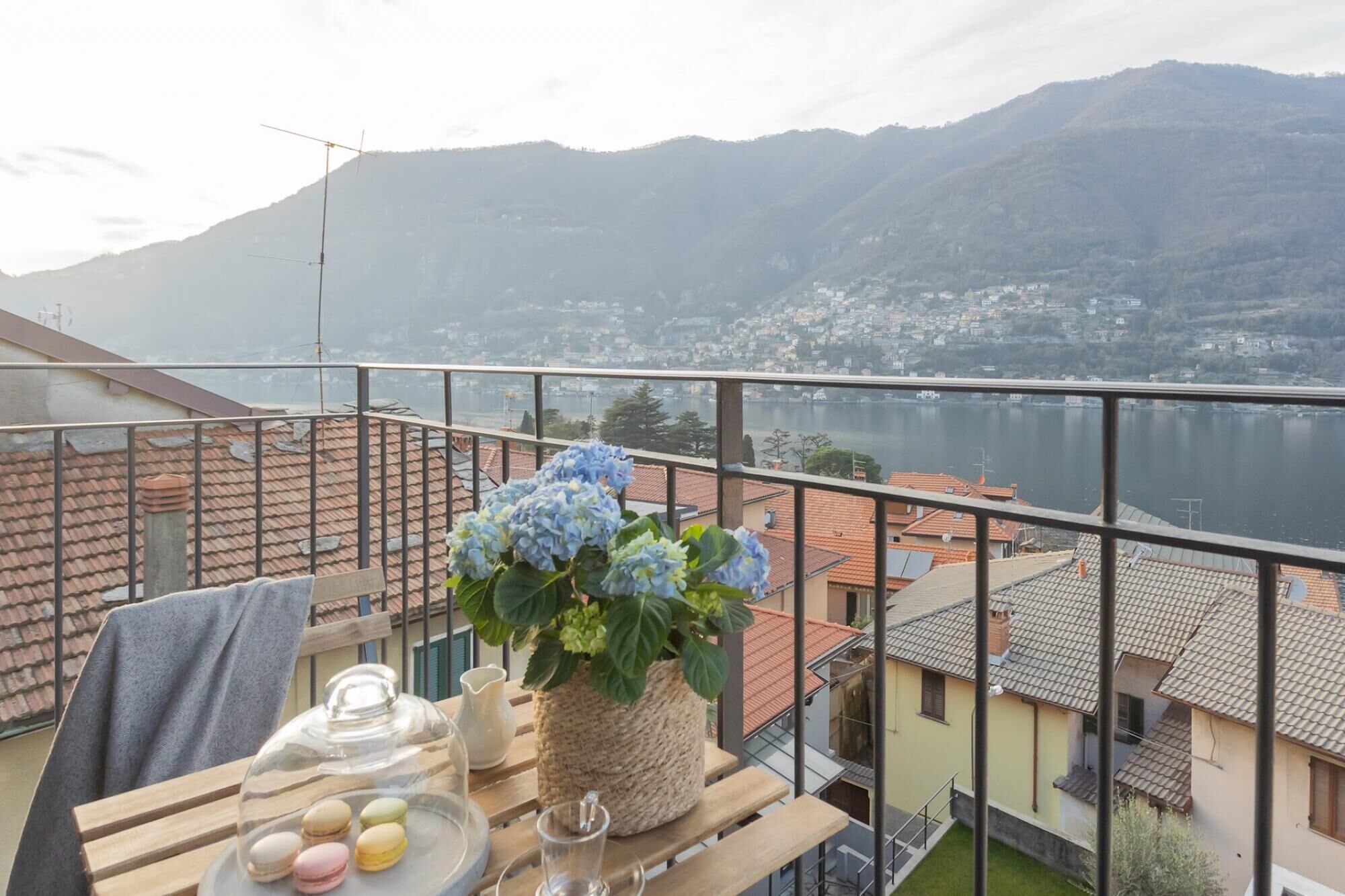 A balcony with a view of a lake and mountains, a wooden table with a vase of flowers, and a glass jar with macarons.