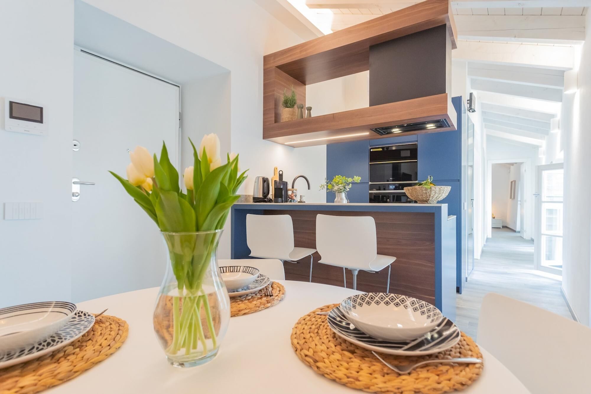 A modern kitchen with a blue countertop, white chairs, and a vase with flowers on the dining table.