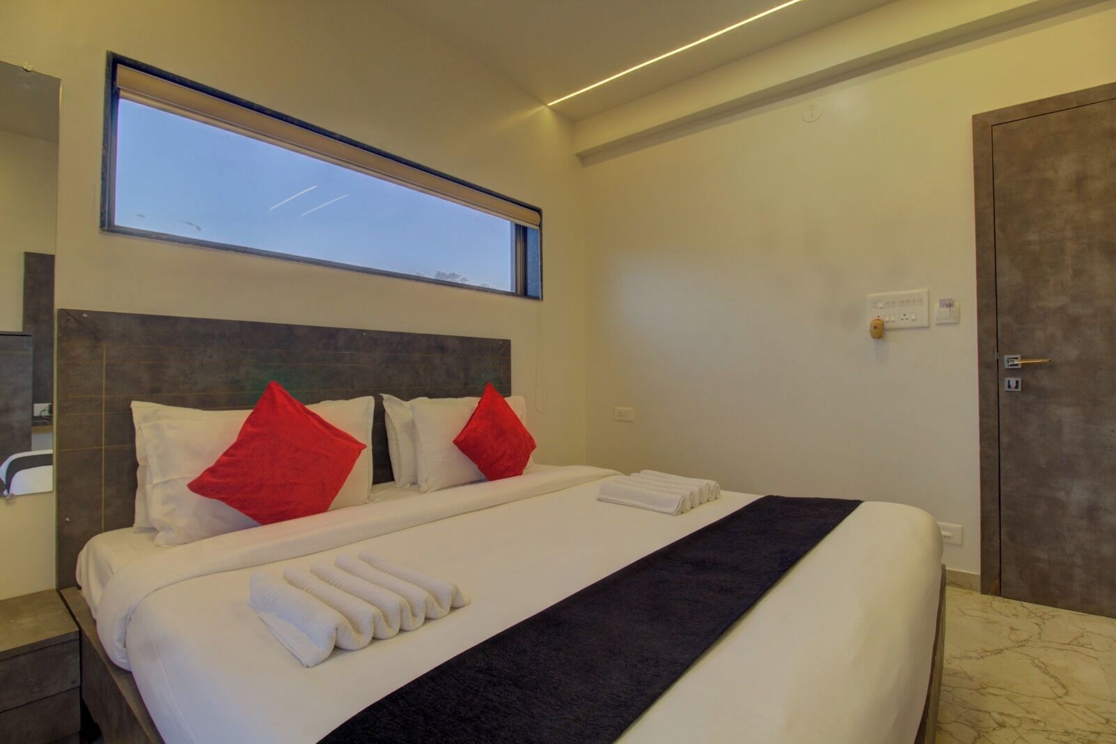 A hotel room with a large bed, two red pillows, a dark blue throw, and a neatly folded white towel.
