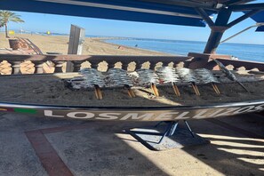 A beachside barbecue grill with fish skewers, a wooden bench, and a view of the beach and ocean.