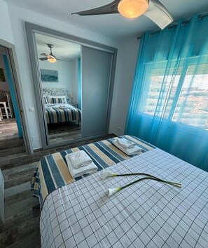A hotel room with a bed, a ceiling fan, a window with curtains, and a view of another room.