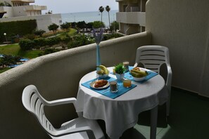 Ein Balkon mit Frühstückstisch, ausgestattet mit Croissants, Obst und einem Getränk, mit Blick auf eine Küstenlandschaft mit Gebäuden und Grünflächen.