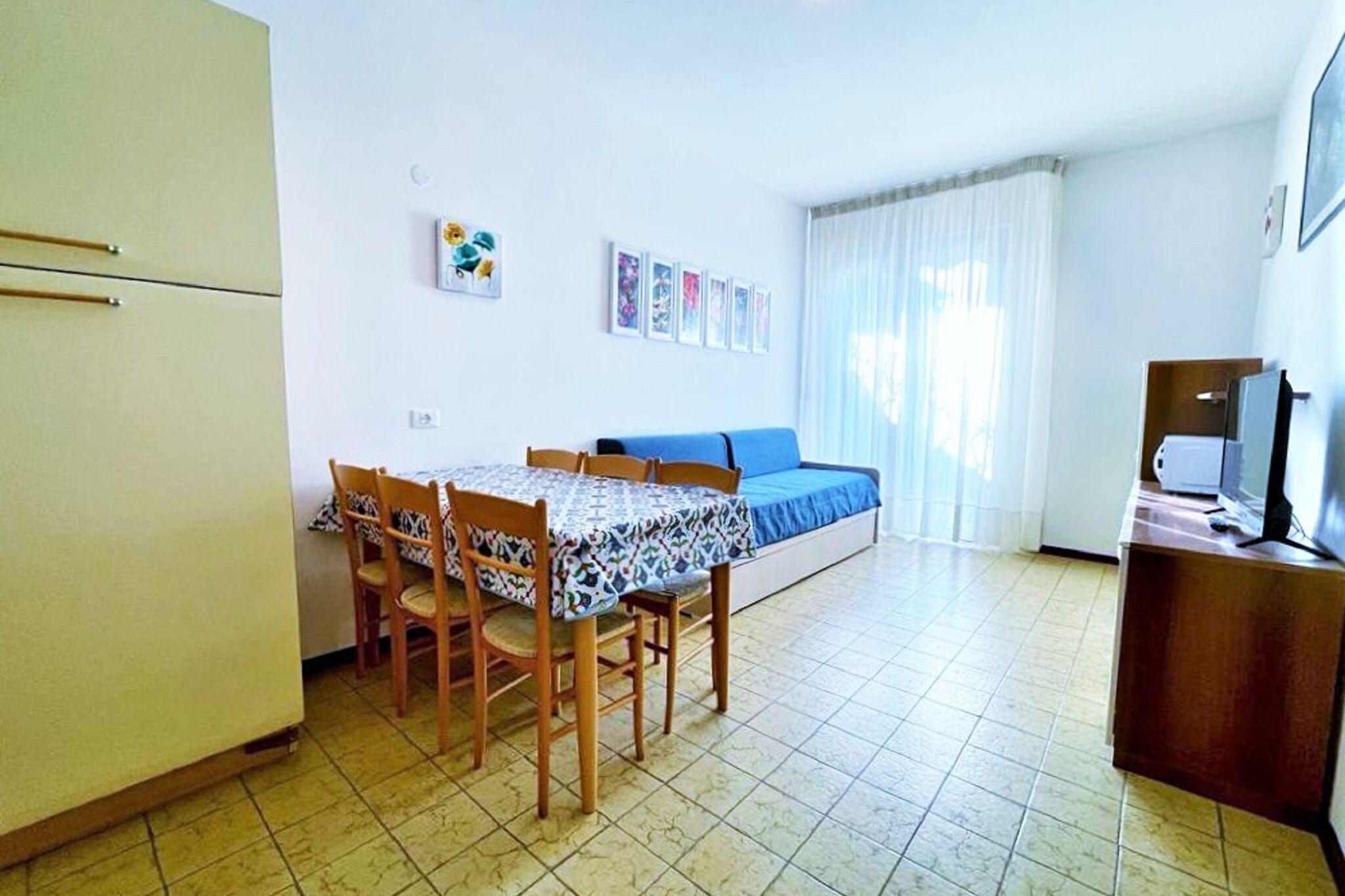 Une petite pièce bien éclairée, équipée d’un lit, d’une table avec des chaises, d’un téléviseur et d’un réfrigérateur.