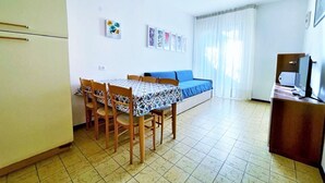 Una habitación pequeña y bien iluminada, con cama, una mesa con sillas, televisor y refrigerador.