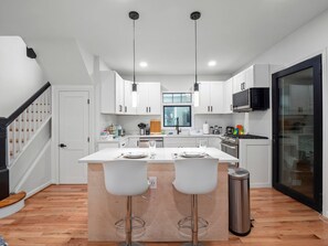 A modern kitchen with white cabinets, a central island with bar stools, stainless steel appliances, and pendant lighting. - Colorful Large City Gatherings (Arlington)