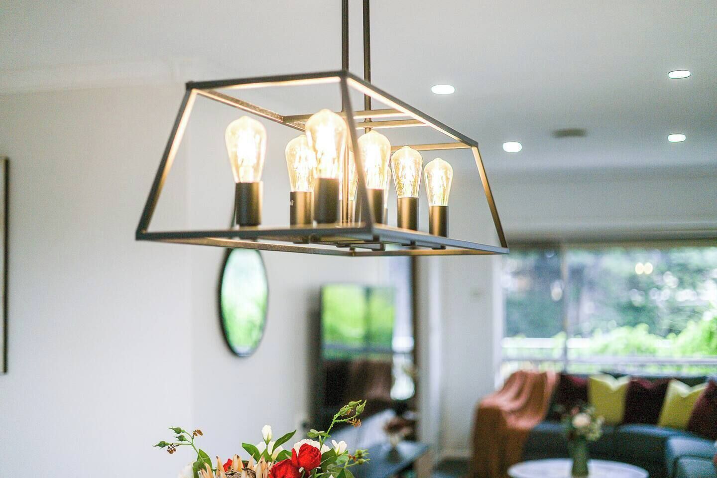 A modern interior with a geometric pendant light fixture with multiple bulbs, a round mirror, and a view of a green outdoor area through large windows.