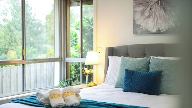 Una cama bien hecha con un edredón verde aguamarina, almohadas blancas y una toalla doblada con la palabra 'Stay' escrita. Hay una lámpara en una mesita de noche y un cuadro en la pared.