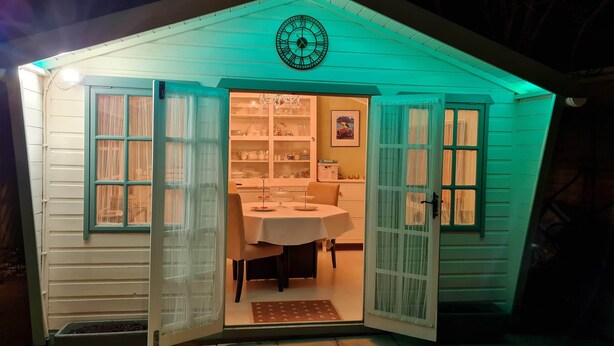 Ein gemütliches, gut beleuchtetes Gartenhaus mit einer Uhr an der Wand, einem Tisch mit Stühlen und einer Tür, die nach innen führt.