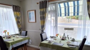 Ein Essbereich mit zwei Tischen, gedeckt für das Frühstück, jeweils mit einer grünen Tischdecke, weißen Tellern und Besteck. Stühle stehen um die Tische, und ein Fenster bietet Blick auf die Straße, wo ein Schild „VACANCIES“ steht.