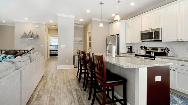 A modern kitchen with white cabinets, a stainless steel refrigerator, a microwave, and a built-in island with bar stools. - The Starfish: Near Huntington Beach State Park (Pawleys Island)
