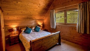 A wooden cabin bedroom with a bed, bedside lamps, and a window with curtains. - The New Farmhouse (Biggar)