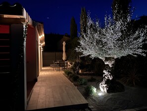 Un patio con zona de comedor, un árbol iluminado con luces y un sendero que lleva a un edificio.