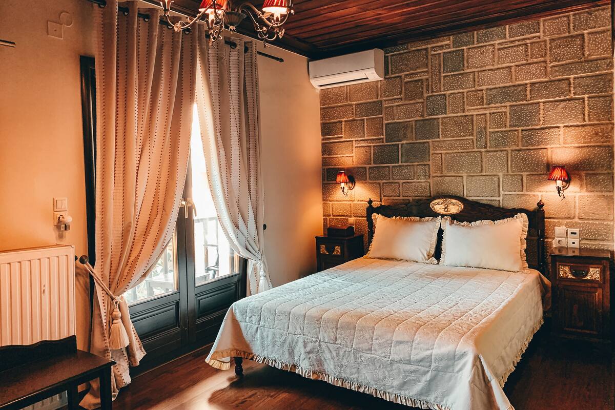 Un dormitorio con cabecera de madera, colcha acolchada, banco de madera, ventana con cortinas y pared de ladrillo.