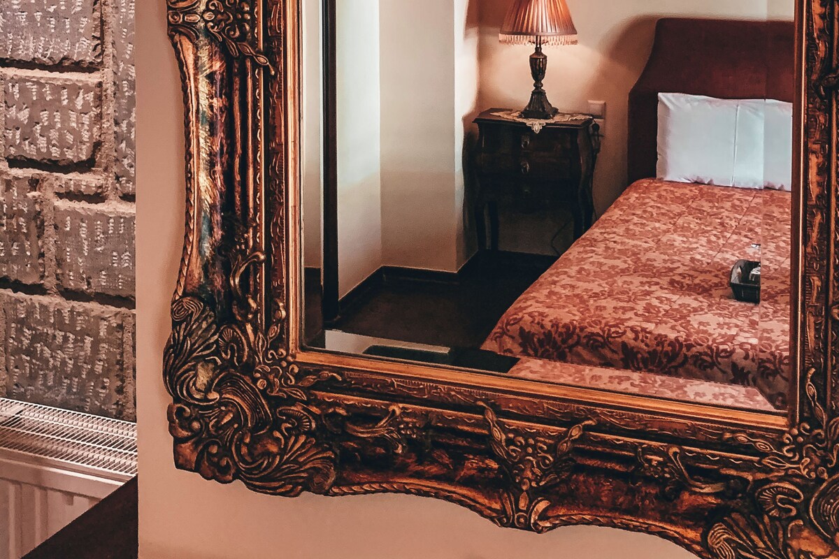 A mirror with an ornate frame reflecting a room with a bed, a lamp, and a brick wall.