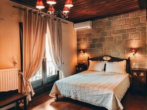 A bedroom with a wooden ceiling, a brick-patterned wall, a bed with a white quilt, and a chandelier. - Guesthouse Elena (Kalabaka)