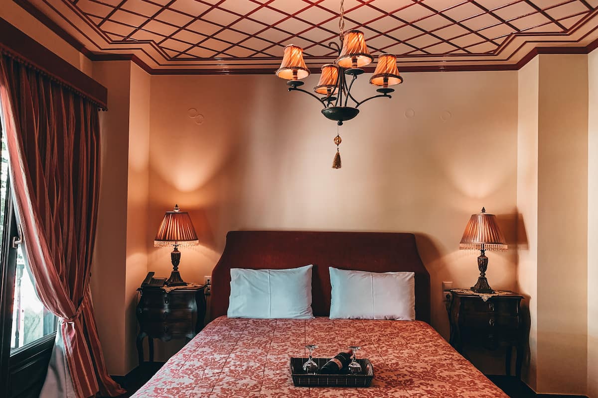 Una habitación de hotel con una cama grande, dos mesitas de noche, un candelabro y un ventanal con cortinas.