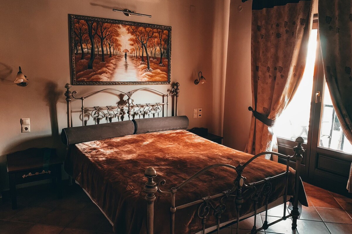 A bedroom with a metal bed frame, a painting of a forest path, a window with curtains, and a wall-mounted air conditioner.