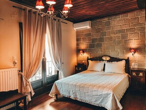 A bedroom with a wooden headboard, a quilted bedspread, a wooden bench, a window with curtains, and a brick wall. - Guesthouse Elena (Kalabaka)