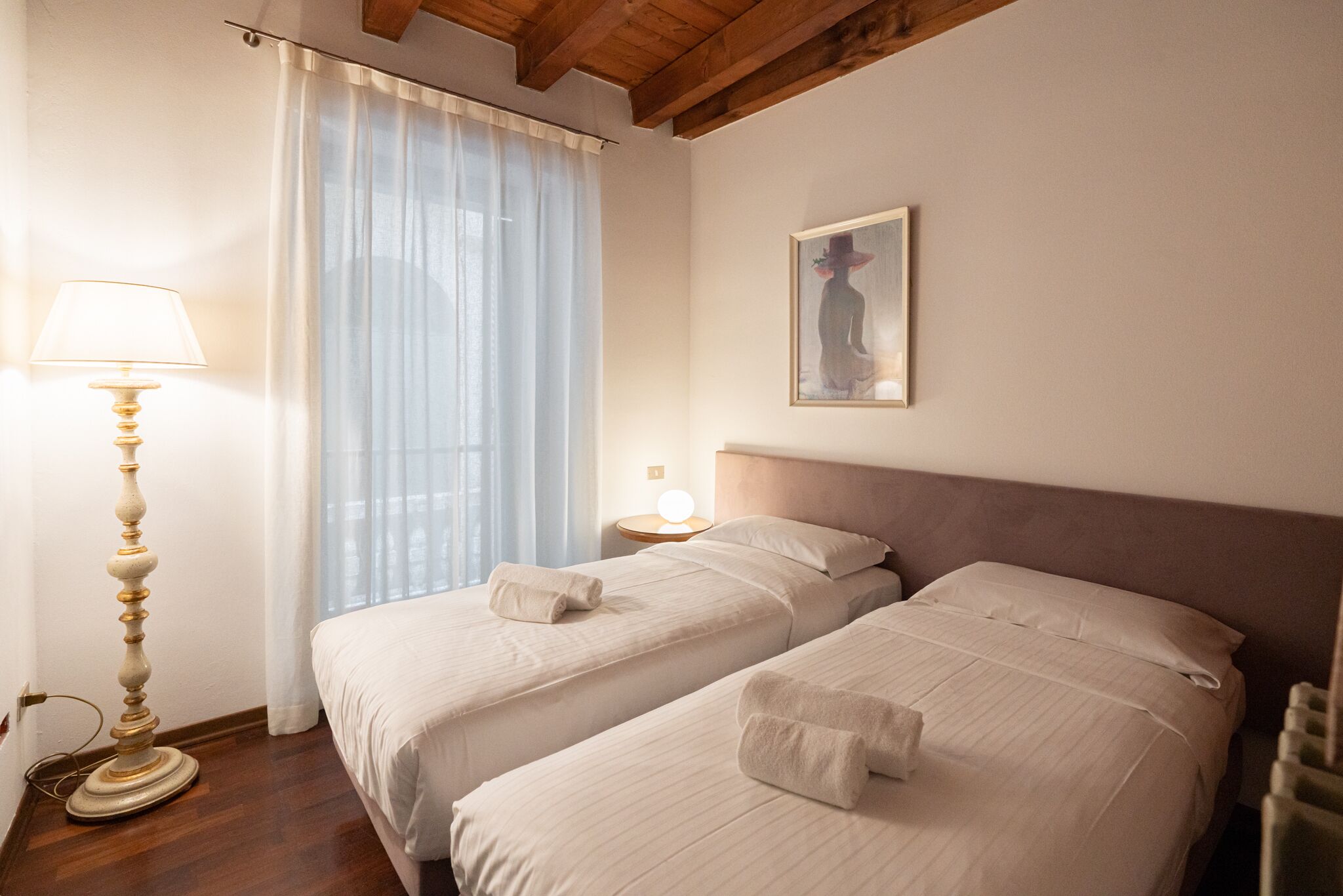Een hotelkamer met twee bedden, een lamp, een schilderij en een raam met gordijnen.