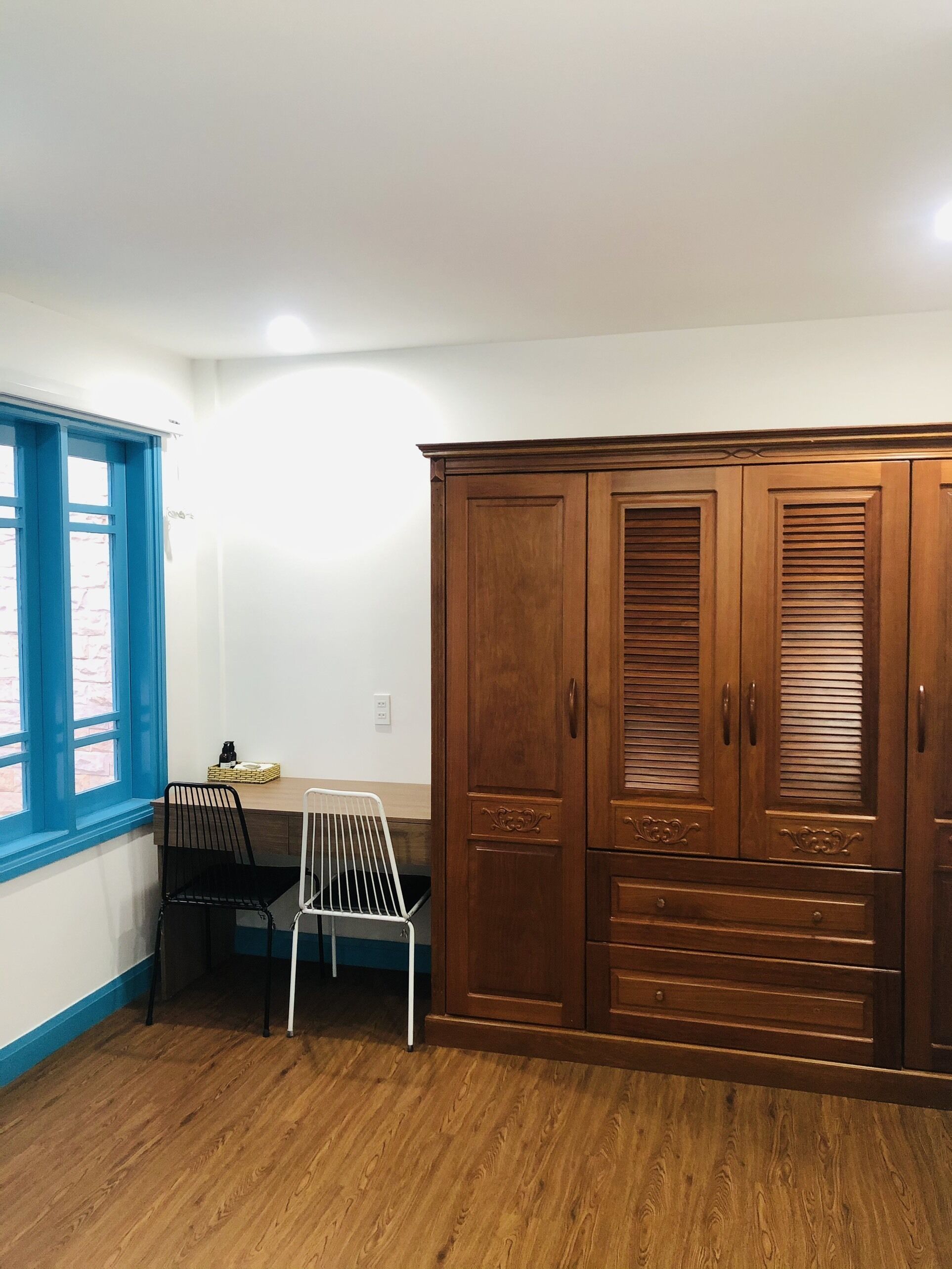Una habitación con un armario de madera, un ventanal con marco azul, una silla blanca y una pequeña mesa con una cesta.